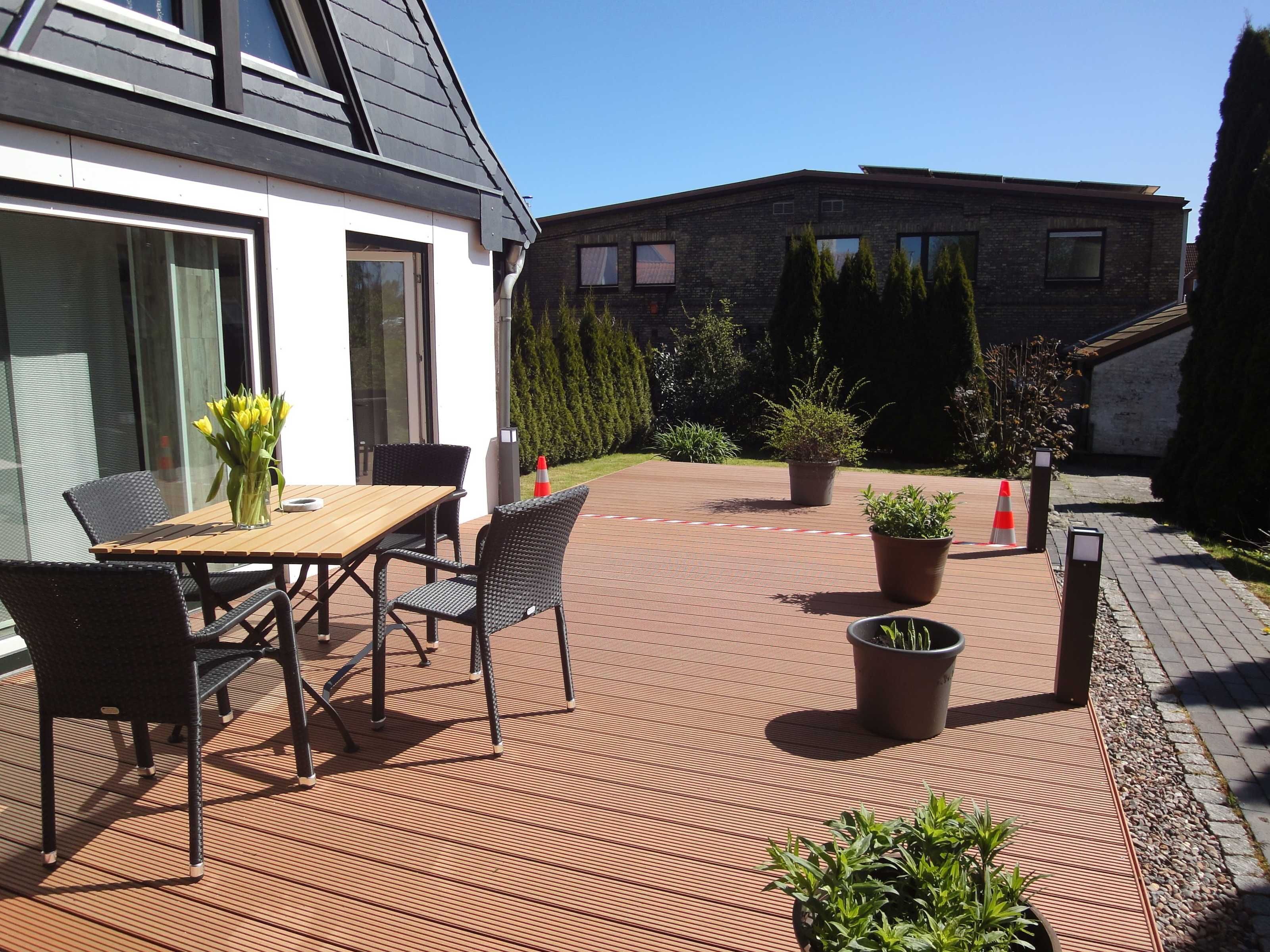 Een terras buiten, met een houten tafel en stoelen, omgeven door plantenpotten en een geplaveid pad.