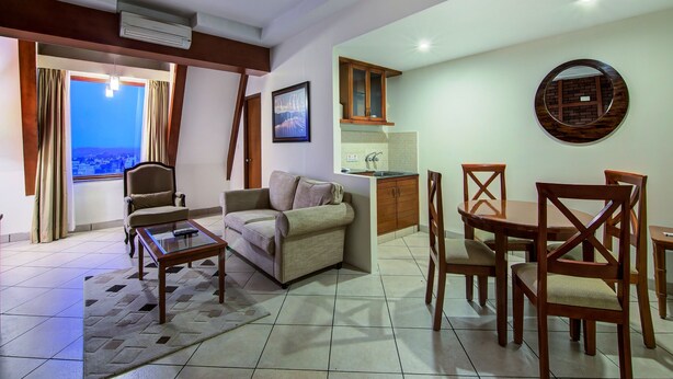 A living room with a sofa, coffee table, and dining area with chairs. There is a kitchen area with a sink and cabinets. A ceiling fan is visible, and a window offers a view of the city. - Sai Vishram Business Hotel (Anekal)