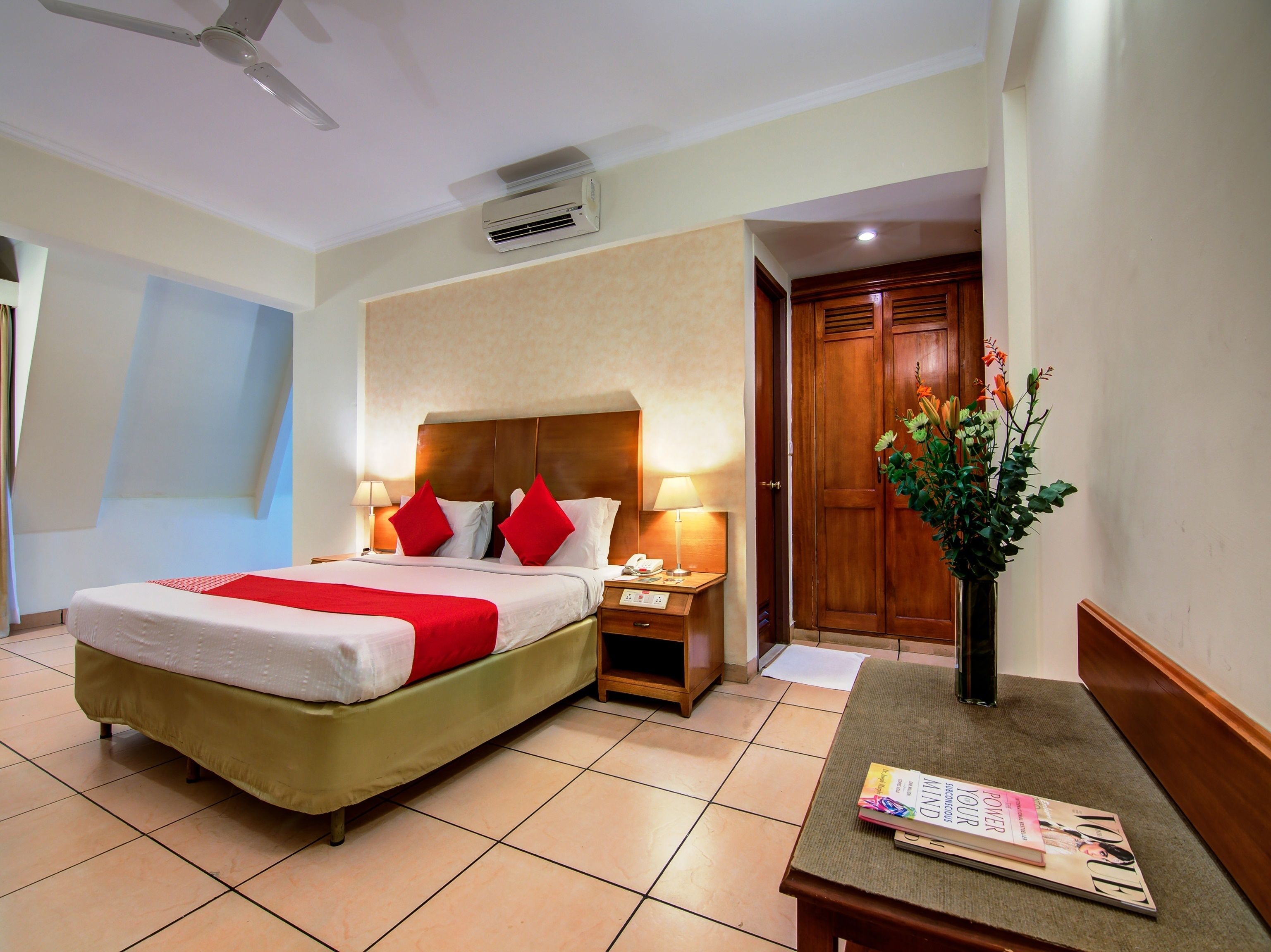 Habitación de hotel con una cama, mesitas de noche, una lámpara, un florero con flores y una revista sobre una mesa.