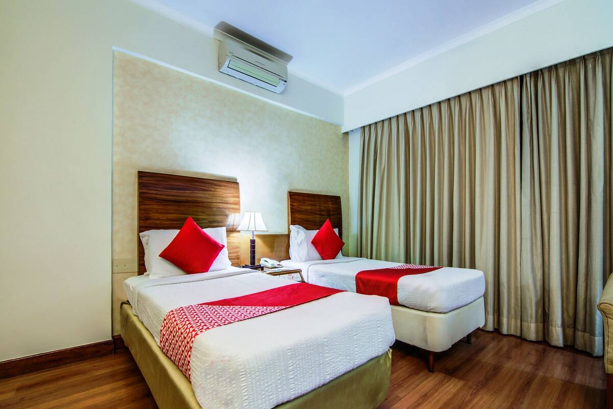 Hotellihuone, jossa on kaksi sänkyä, suuri ikkuna, jossa on verhot, yöpöytä lampun kanssa ja ilmastointilaite.