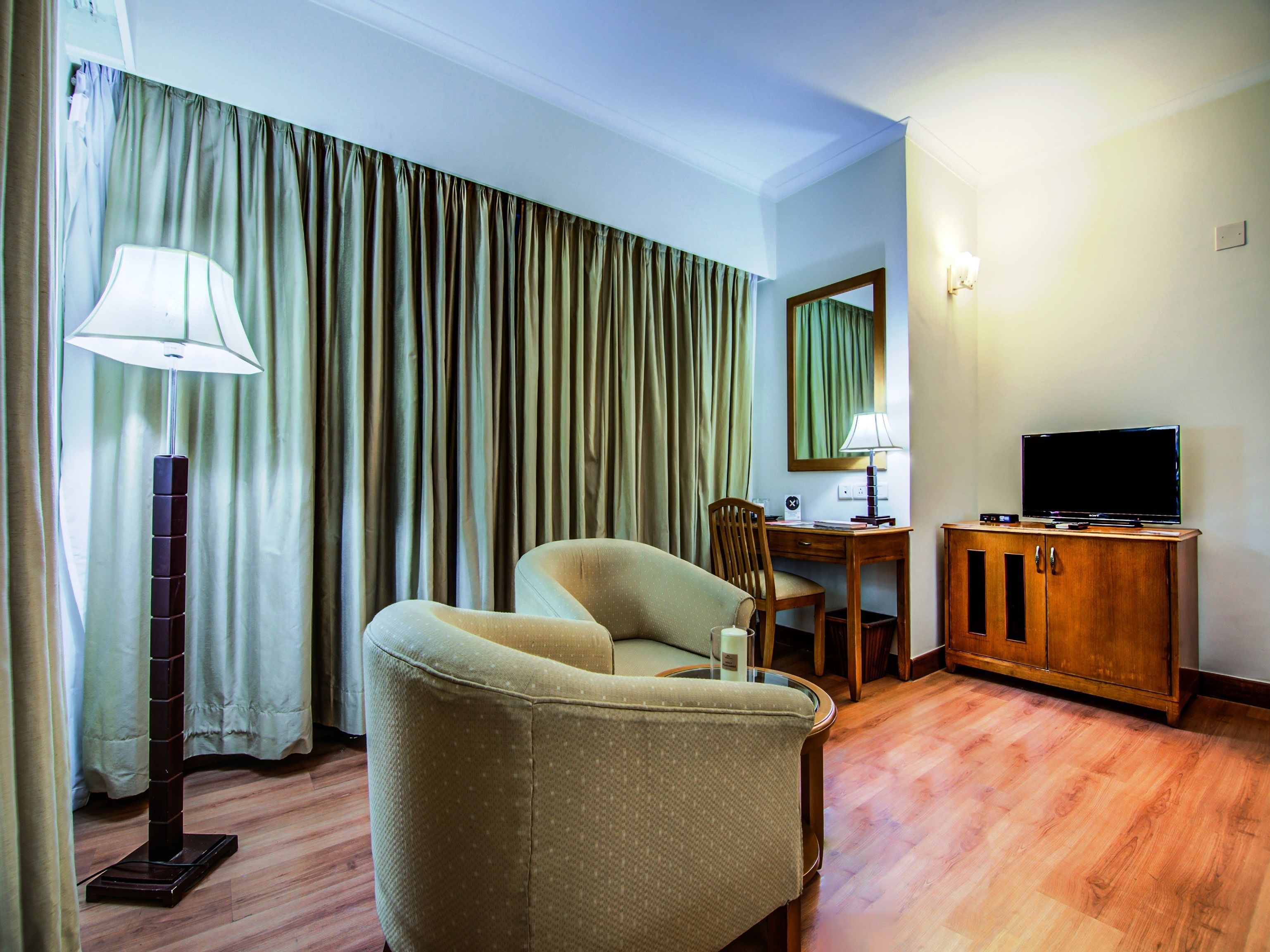 A hotel room with a sofa, a desk with a chair, a television on a wooden cabinet, and a lamp.