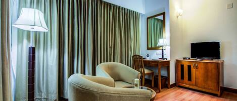 A hotel room with a sofa, a desk with a chair, a television on a wooden cabinet, and a lamp.