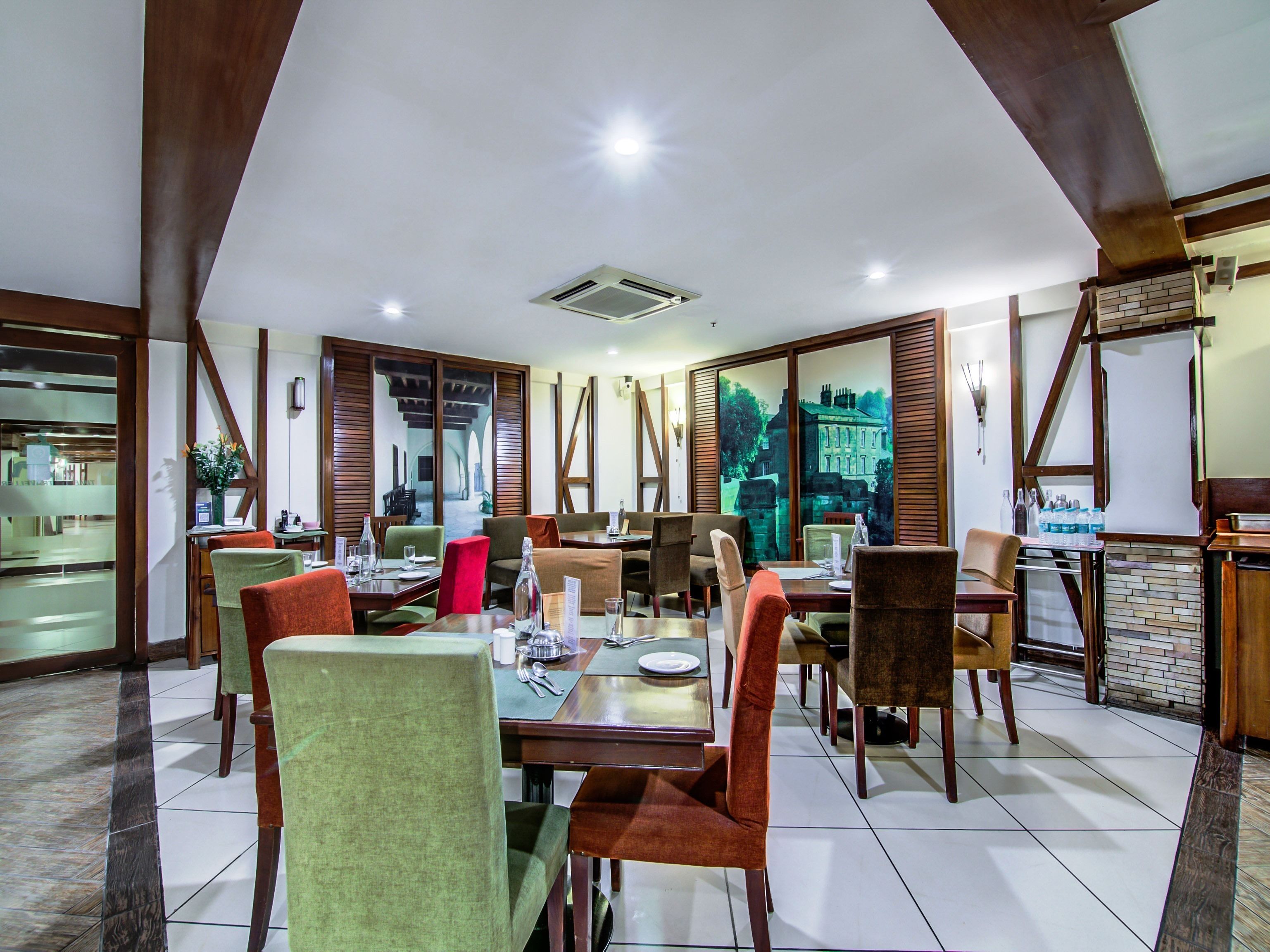 Un restaurante con techo alto, vigas de madera y un ventanal con vistas a un edificio histórico. Las mesas están preparadas con platos, vasos y cubiertos.
