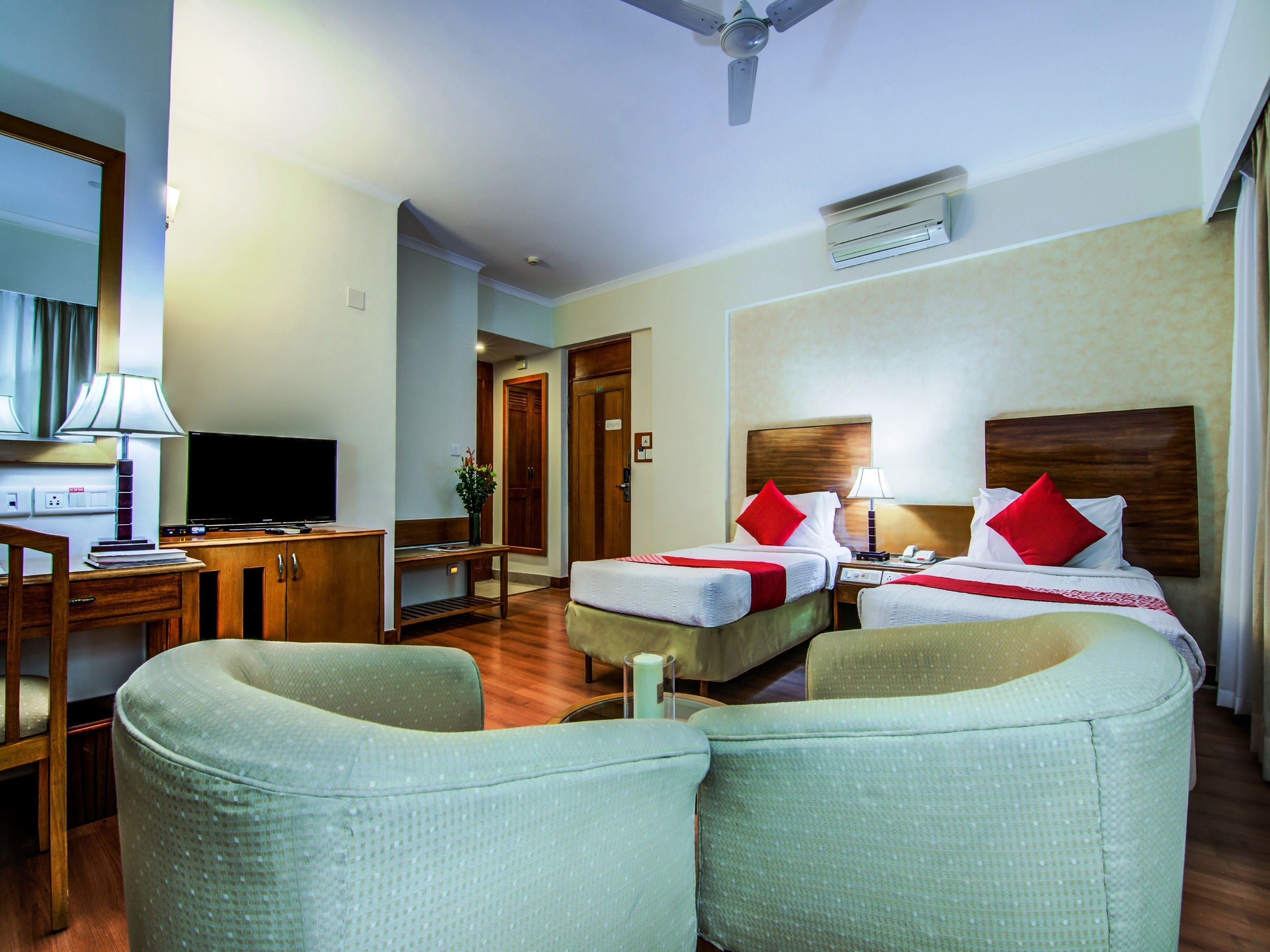 Une chambre d’hôtel avec deux lits, un bureau avec une télévision, une chaise et un ventilateur de plafond.