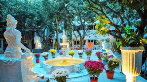 Un patio con una fuente central, rodeado de macetas con plantas e estatuas iluminadas.