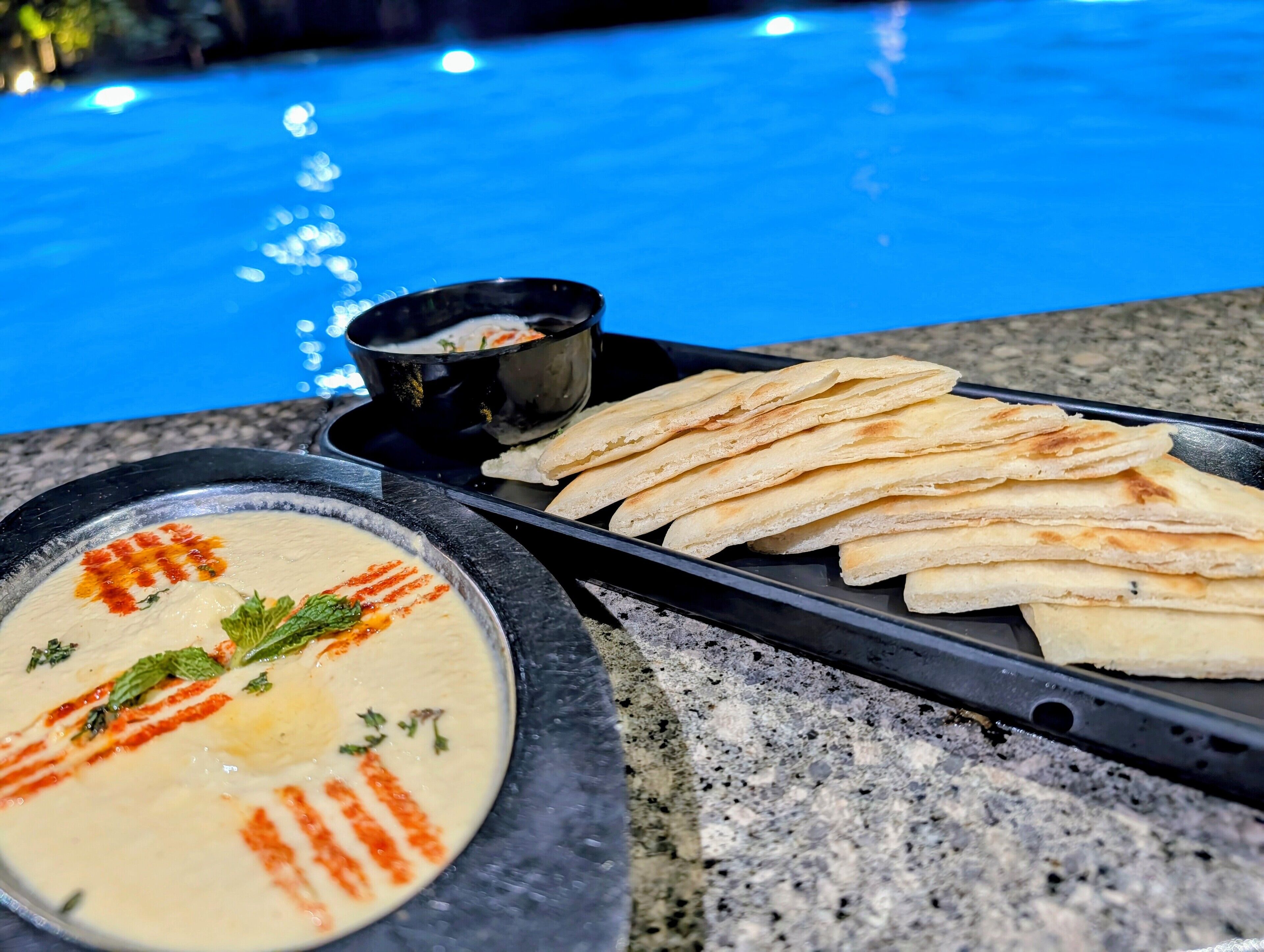 A dining setup with a bowl of soup garnished with herbs and a small bowl of sauce, accompanied by flatbreads on a tray.