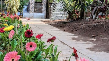 Een tuinpad met roze en gele bloemen, een klein prieeltje en een trap die naar een gebouw leidt.