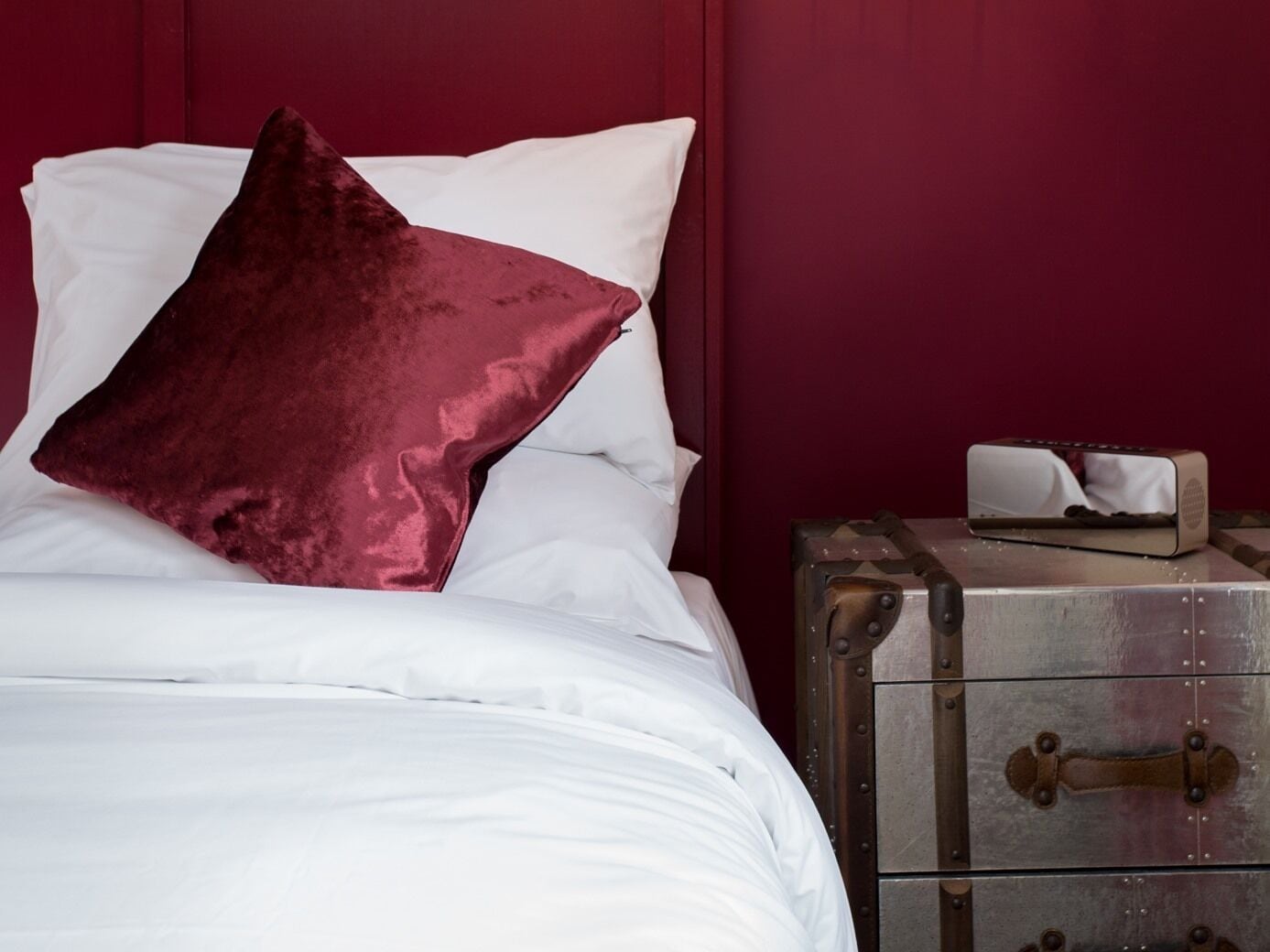 Een bed met witte lakens en een donkerrode kussen, een vintage koffer op een metalen dressoir, en een klein voorwerp op het dressoir.