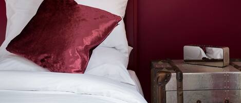Una cama con sábanas blancas y una única almohada rojo oscuro, una maleta antigua sobre un tocador de metal y un pequeño objeto en el tocador.