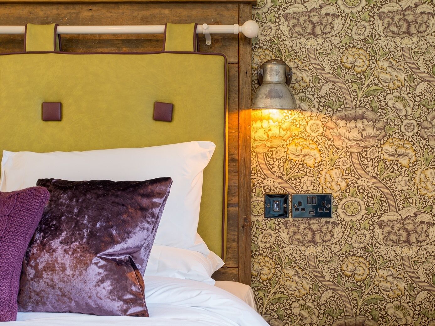 Un lit avec une tête de lit verte, des oreillers blancs et violets, et un luminaire fixé au mur.