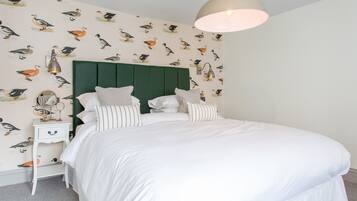 Un dormitorio con cabecera verde, ropa de cama blanca, almohadas a rayas, una mesita de noche con espejo y un papel tapiz con motivos de pájaros.
