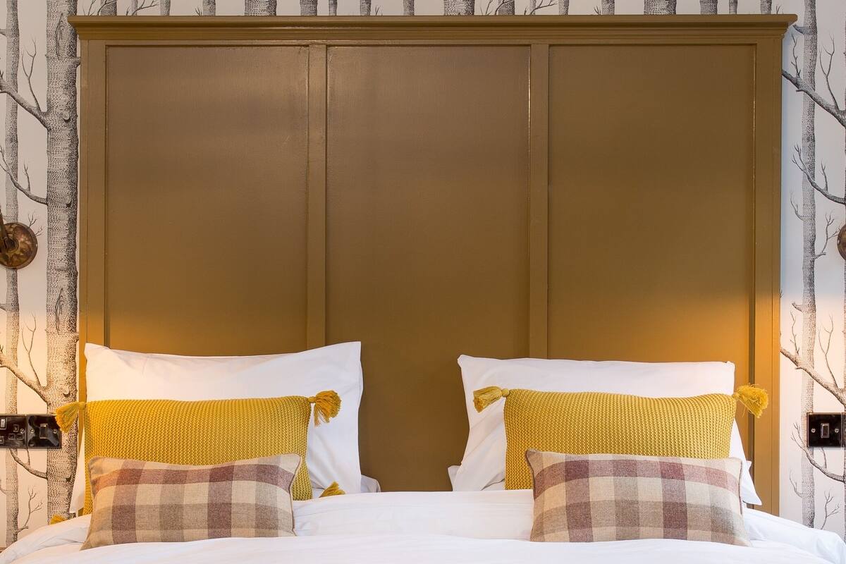Una cama con sábanas blancas y dos almohadas amarillas, cabecera marrón y pared con estampado de árbol.