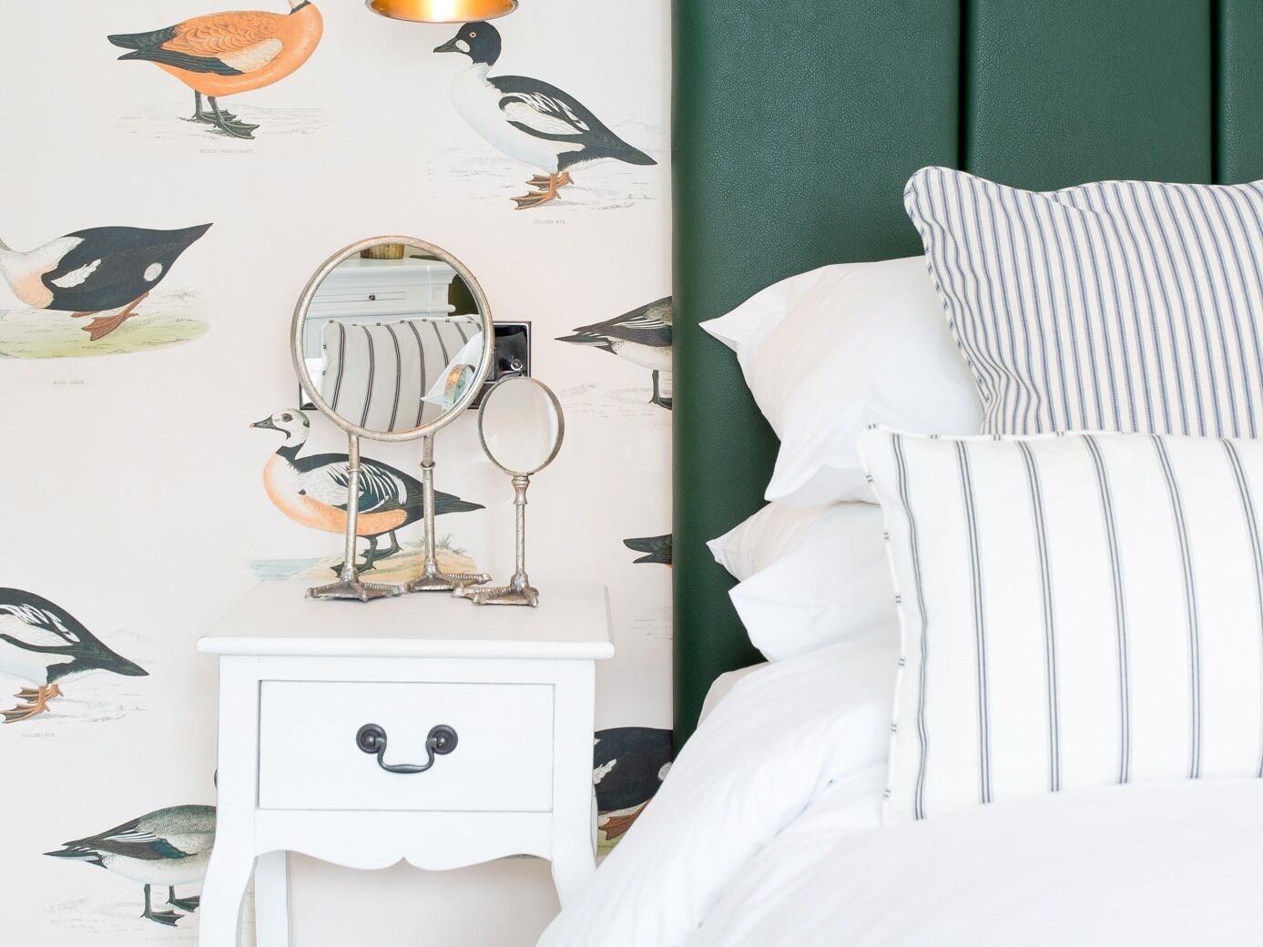 A bedroom with a white nightstand, a mirror, and a drawer with a face design. The wall has a bird-themed wallpaper. There is a bed with white bedding and striped pillows.