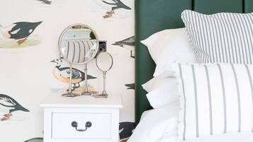 A bedroom with a white nightstand, a mirror, and a drawer with a face design. The wall has a bird-themed wallpaper. There is a bed with white bedding and striped pillows.