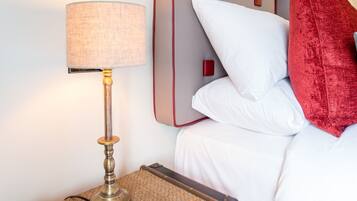 A neatly made bed with white bedding and a red pillow, a lamp on a woven side table, and a suitcase on the floor.