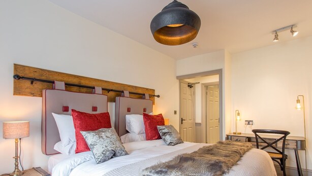 A double bed with a large fur throw, a wooden headboard, and red and grey pillows. There is a desk with a chair, a lamp, and a door in the background. - The Cornish Arms (Tavistock)