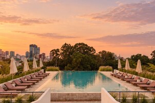 Una piscina rectangular con sillones de descanso y sombrillas, rodeada de follaje y un horizonte urbano al atardecer.