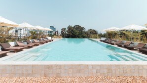 A rectangular swimming pool with clear blue water, surrounded by lounge chairs and umbrellas, set against a backdrop of buildings and trees. - Penh House Hotel (Phnom Penh)