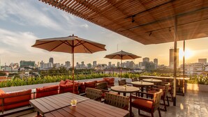 An outdoor seating area with tables and chairs, large umbrellas, and a city skyline view in the background. - Penh House Hotel (Phnom Penh)