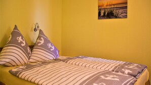 Ein Bett mit gestreifter Bettwäsche, zwei Kissen mit Anker-Motiven, eine Wandlampe und ein Sonnenuntergangsbild an der Wand.
