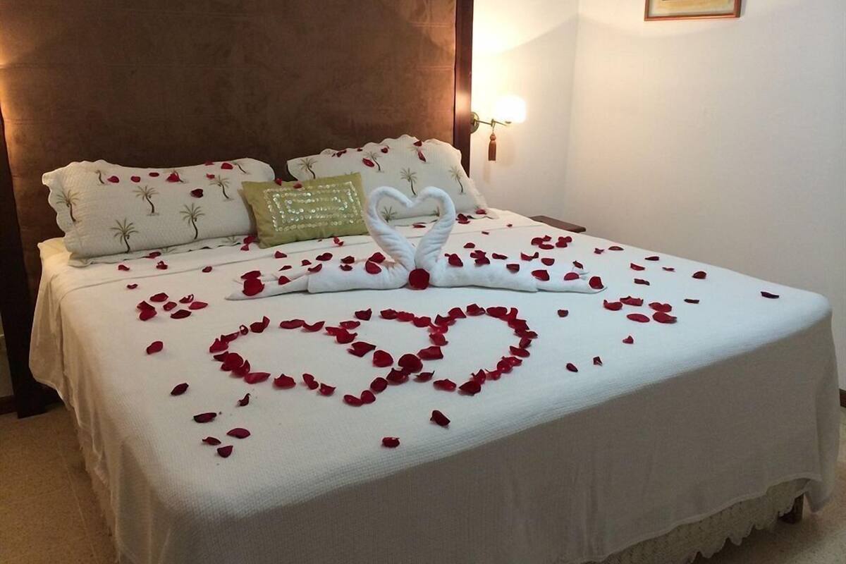 Ein ordentlich bezogenes Bett mit Rosenblättern, die in Herzform drapiert sind, einer Schwan-Dekoration, Kissen und einer Wandleuchte.