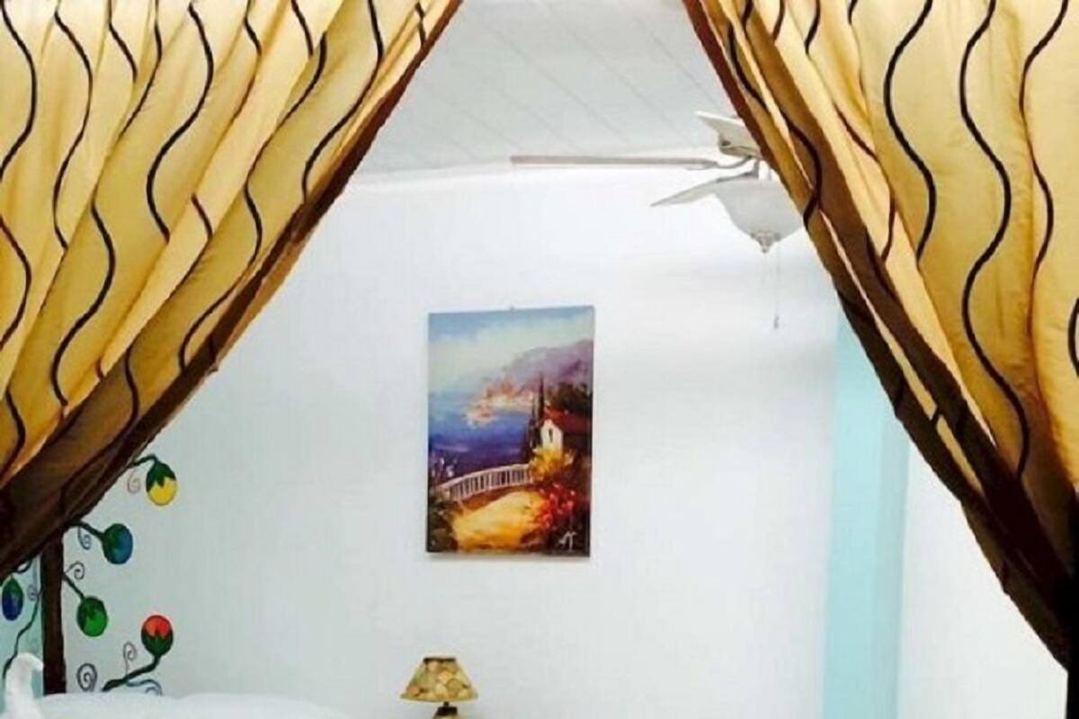 Ein Hotelzimmer mit drei Betten, jedes mit weißer Bettdecke und einem dekorativen Kopfkissen. Darüber an der Wand hängt ein Gemälde, und auf dem Nachttisch steht eine Lampe.
