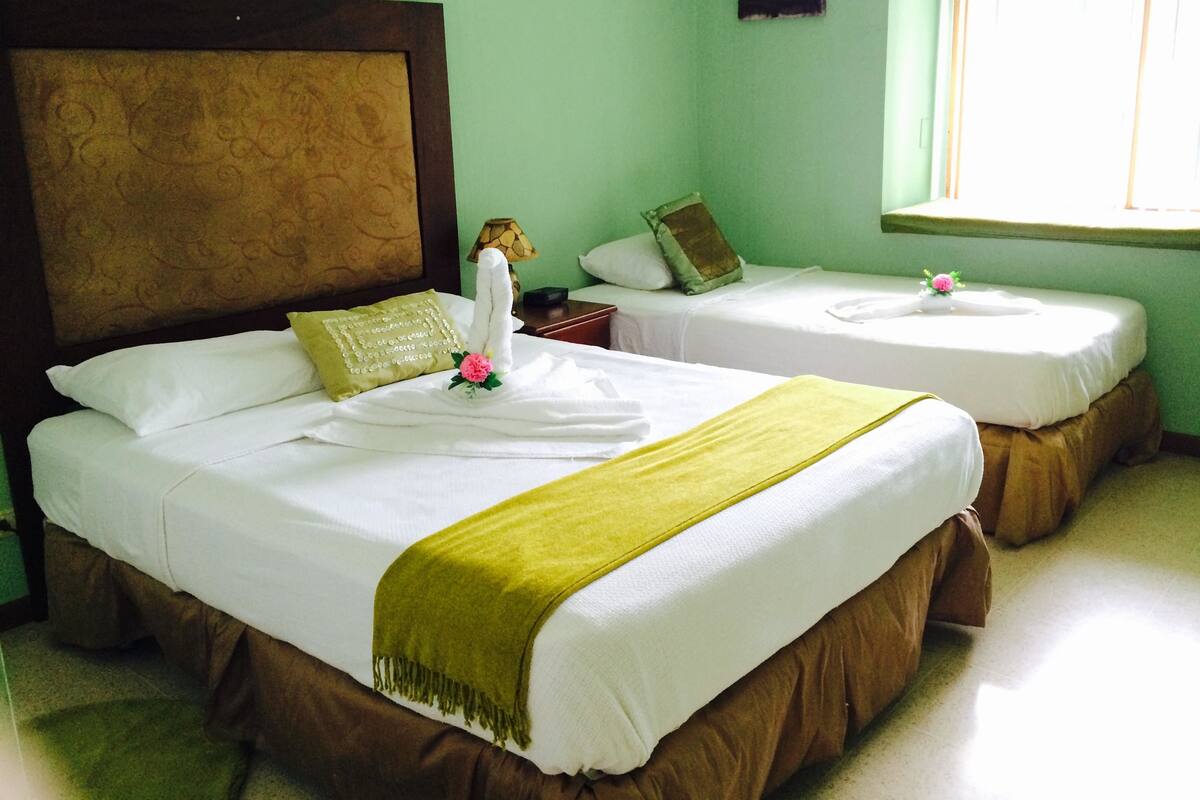 Ein Hotelzimmer mit zwei Betten, beide mit weißen Bettwäsche und einer grünen Tagesdecke. Am linken Bett ist ein Holz-Kopfteil und dazwischen ein Nachttisch mit einer Lampe.