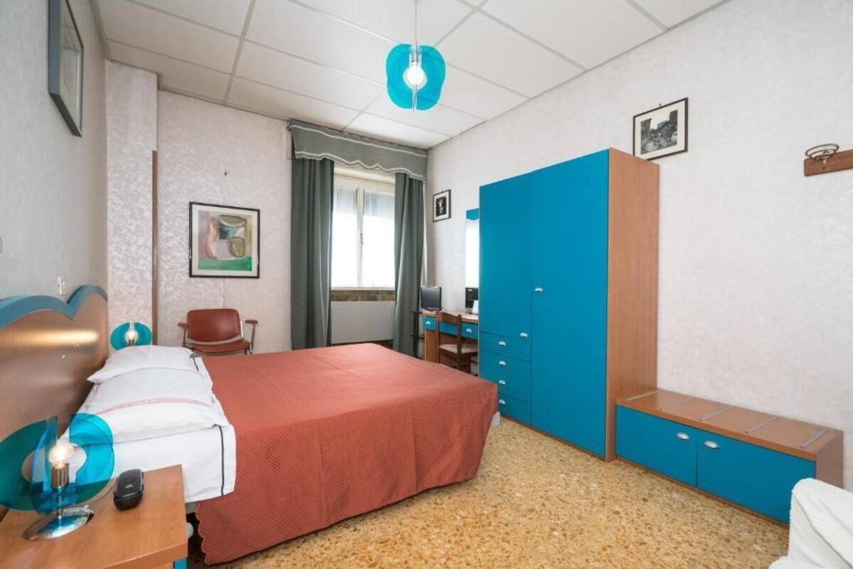 A hotel room with a bed, a desk with a computer, a chair, a wardrobe, and a window with curtains.
