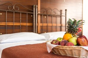 A neatly made bed with a wicker fruit basket containing apples, peaches, a pineapple, and an orange.
