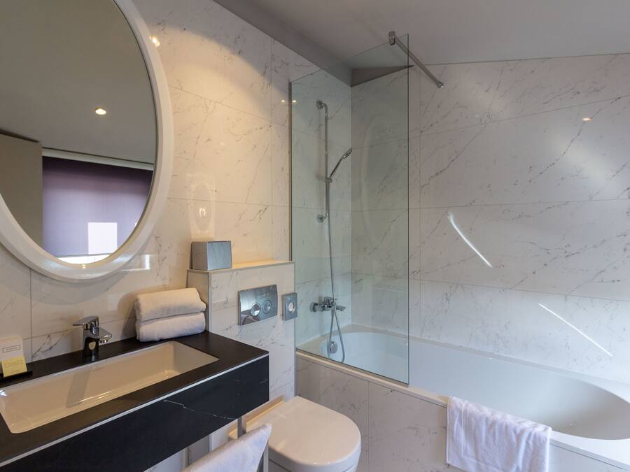 Un baño moderno con un espejo ovalado grande, un lavabo con encimera negra, un inodoro blanco y una cabina de ducha con paredes de vidrio.