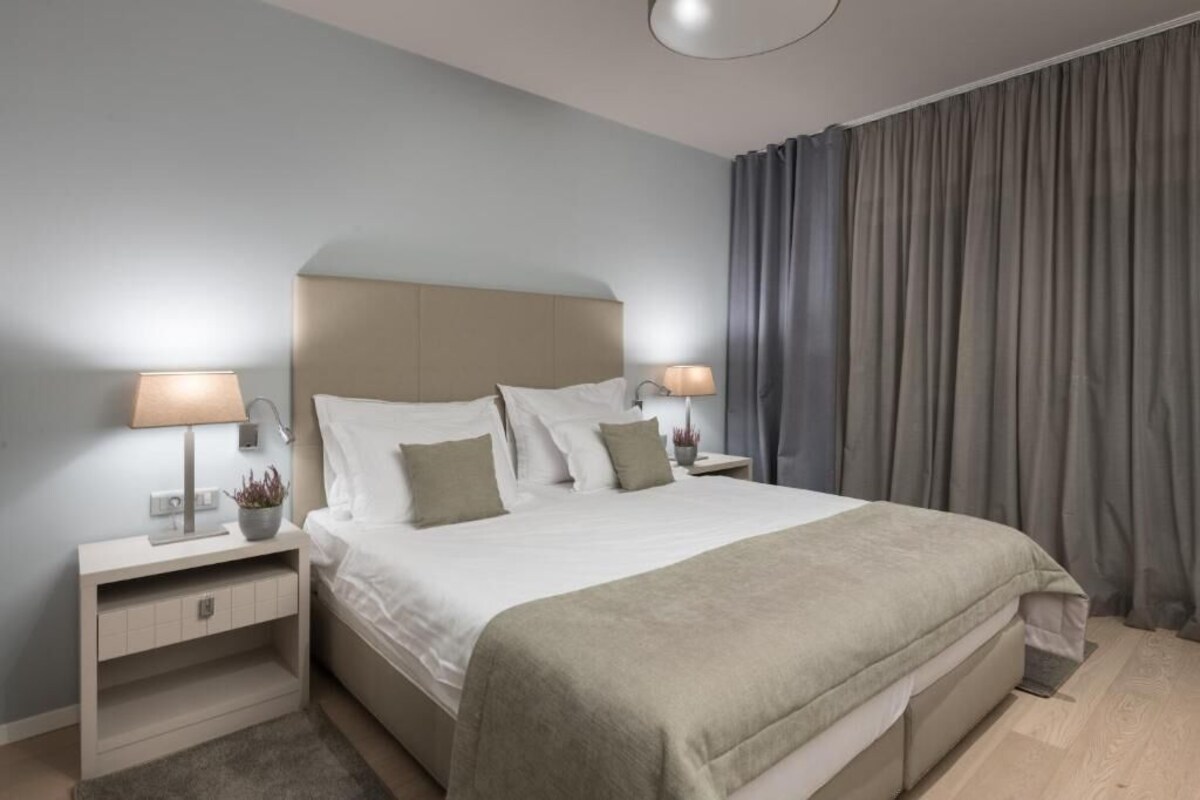 Een moderne hotelkamer met een groot bed, bedlampjes, een nachtkastje, een kleine plant en een raam met gordijnen.