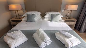 Un lit bien fait, avec du linge de lit blanc et des oreillers gris, deux tables de chevet avec des lampes, et une couverture grise posée sur le sol.
