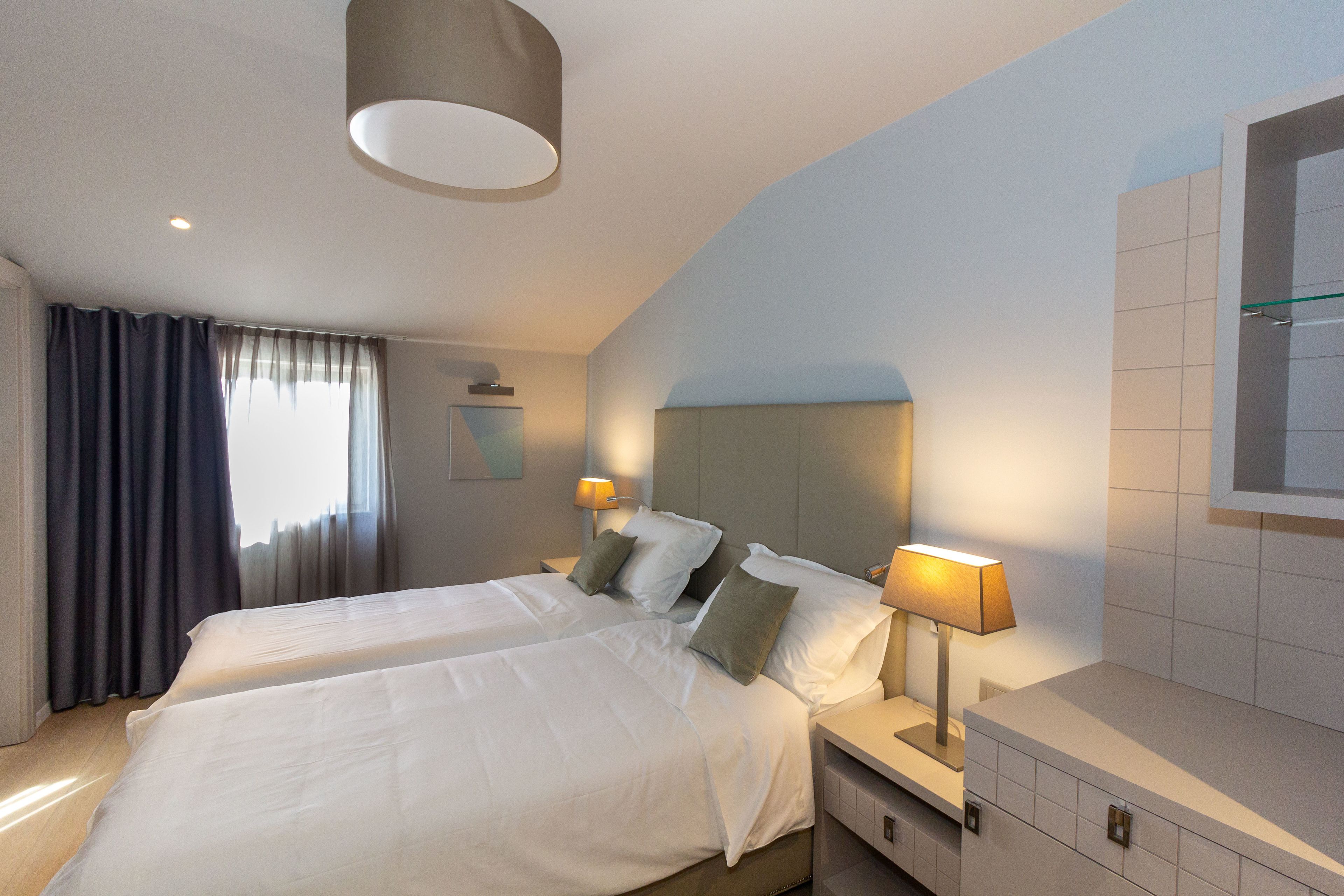Habitación de hotel con dos camas, un ventanal grande con cortinas, un cabecero moderno, lámparas de noche y un televisor integrado.