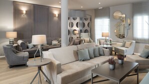 Una sala de estar moderna con un sofá beige grande, dos sillones, una mesa de centro y una mesita auxiliar con una lámpara. Se observan paneles decorativos en la pared y un espejo con diseños circulares.