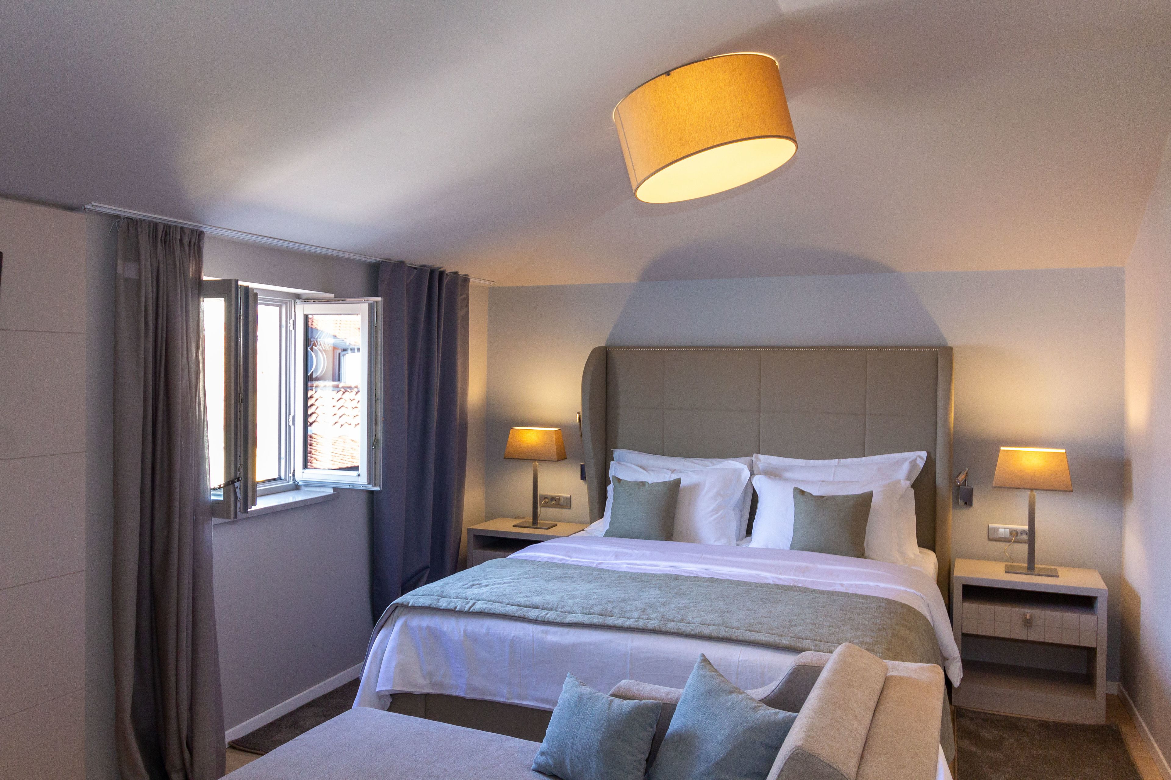 A hotel room with a large bed, two bedside tables with lamps, a window with curtains, and a ceiling light fixture.
