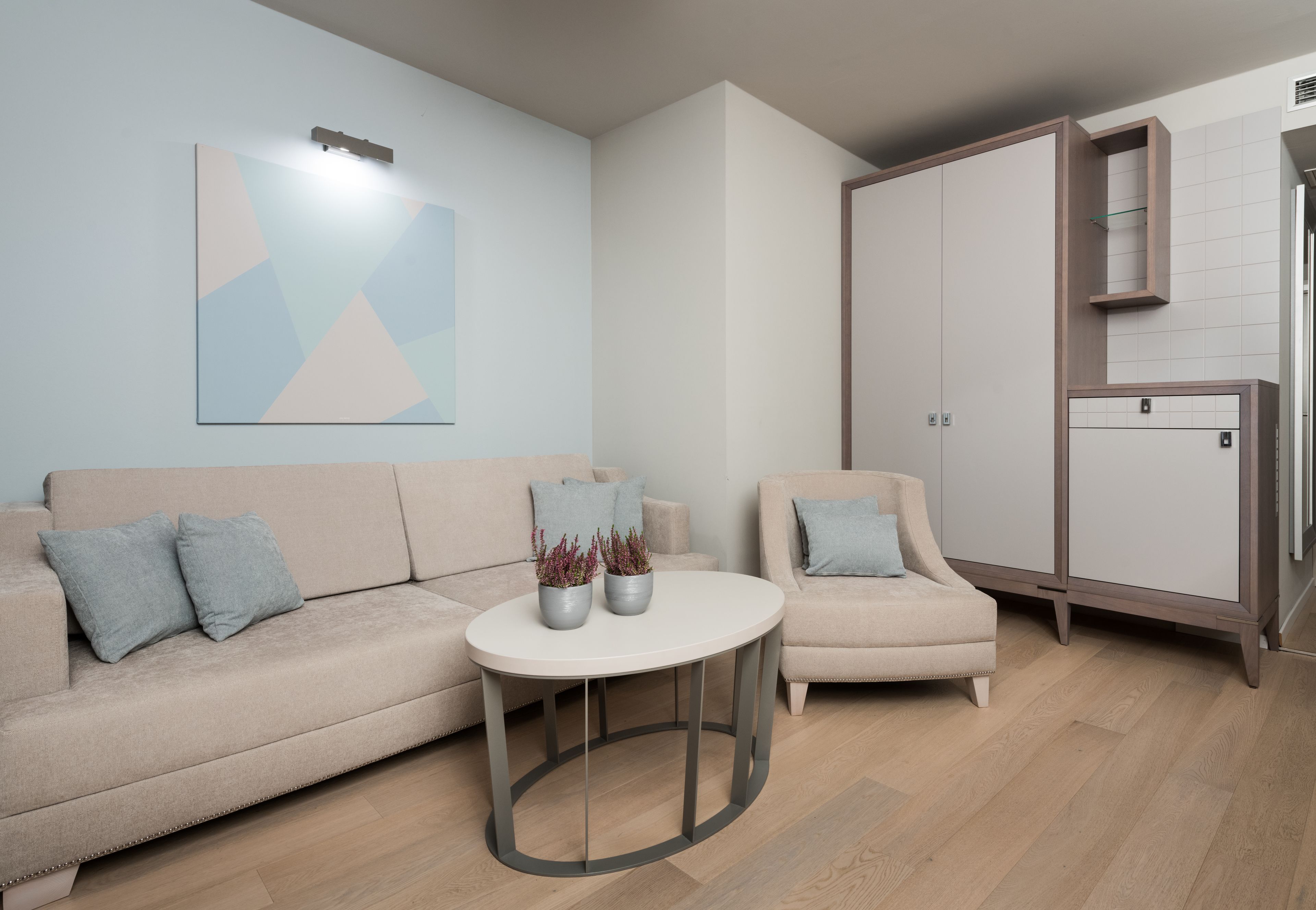 Una sala de estar moderna con un sofá color beige, una mesa de centro redonda y un sillón pequeño. Hay una estantería de pared con objetos decorativos y un cuadro abstracto grande.
