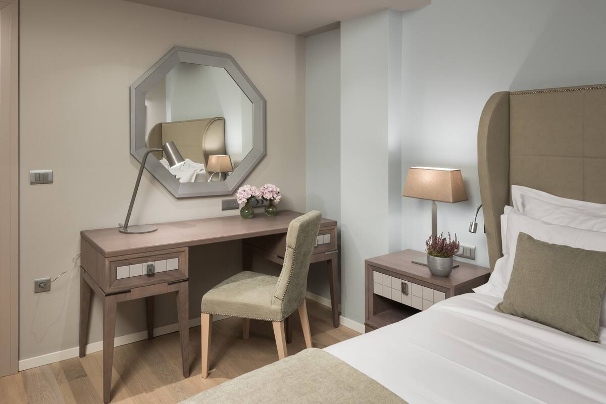 Habitación de hotel con una cama, un escritorio con silla, una mesita de noche con lámpara, un espejo y una lámpara fijada a la pared.