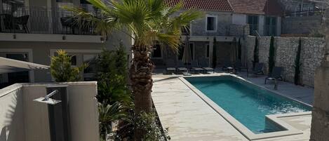 A swimming pool with lounge chairs, a palm tree, and stone buildings in the background.