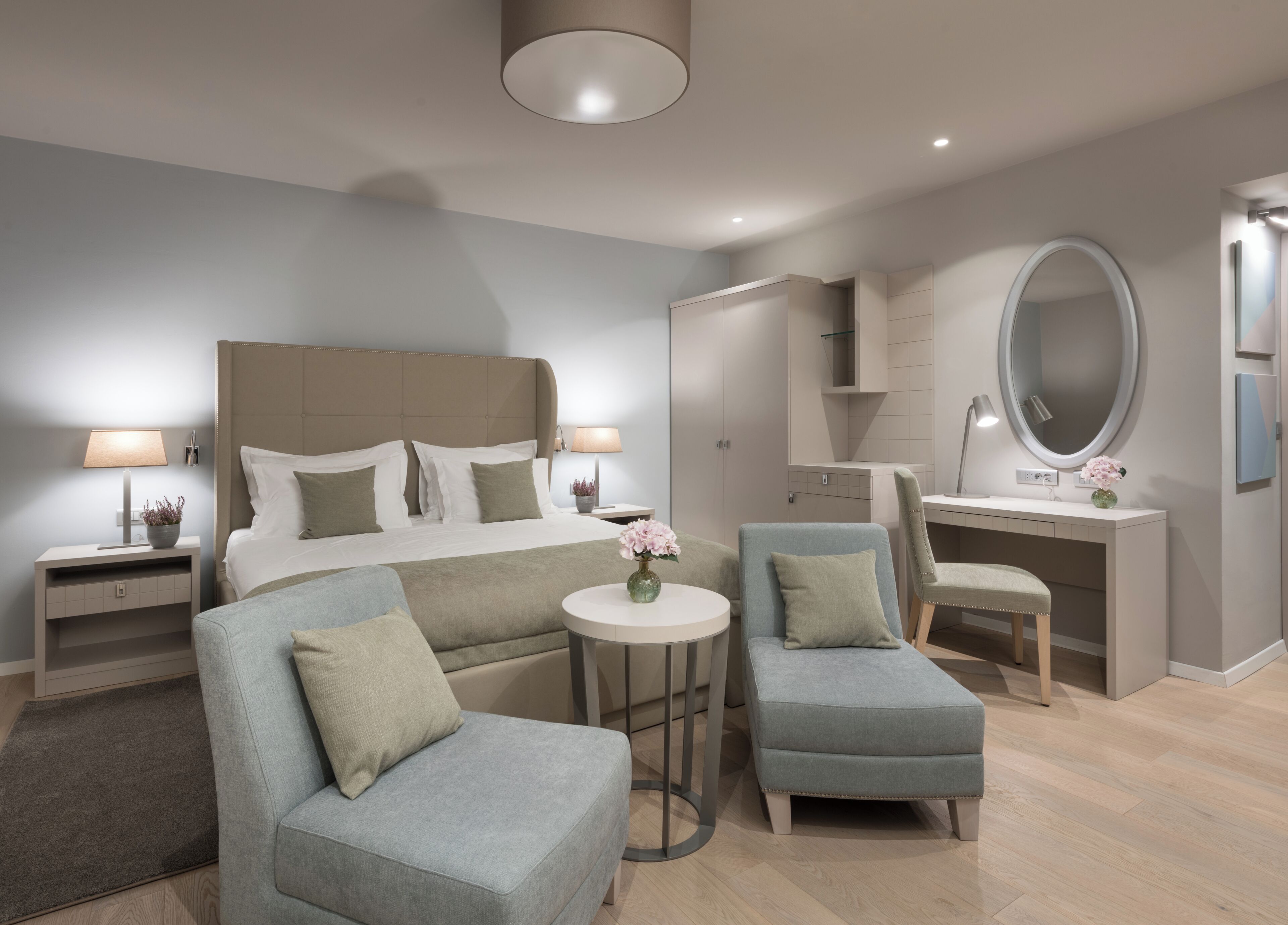 A modern bedroom with a large bed, two armchairs, a desk, and a round mirror.