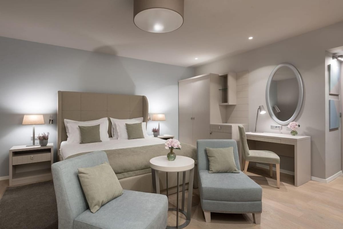 Une chambre d’hôtel moderne comprenant un lit, deux fauteuils, un bureau, un miroir rond et une petite table avec un vase.