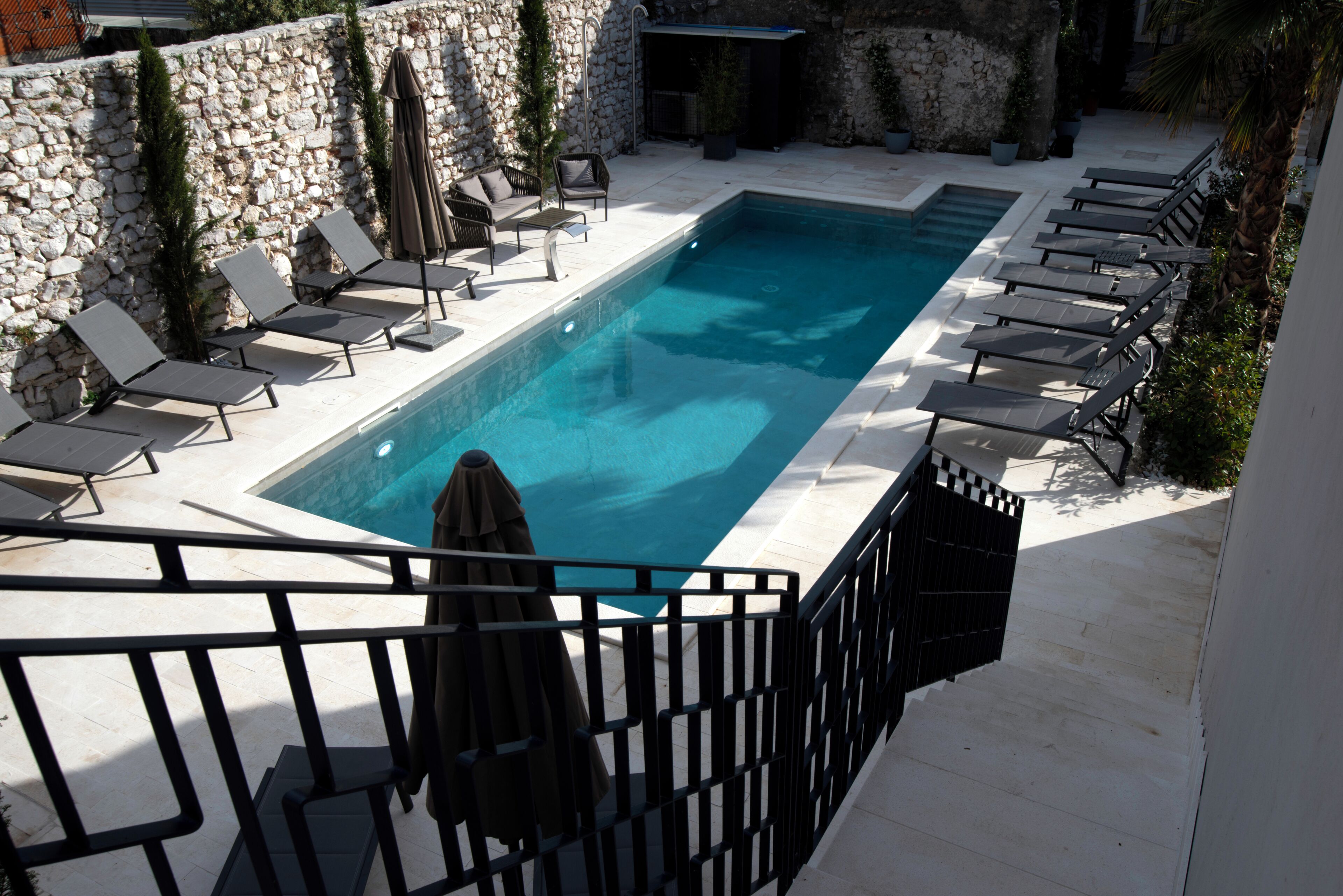 A rectangular swimming pool with lounge chairs and umbrellas, surrounded by stone walls and potted plants.