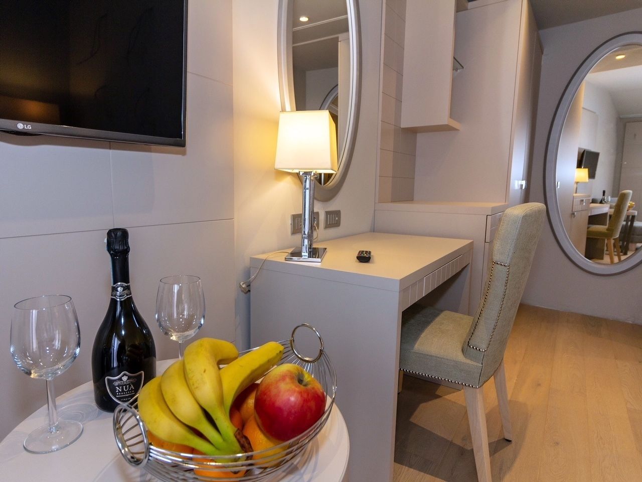A hotel room with a desk, a chair, a TV mounted on the wall, and a table with a fruit bowl, wine glasses, and a bottle.