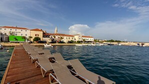 Un muelle de madera con sillones de descanso que se extiende sobre aguas tranquilas, un hotel con una torre al fondo y barcos amarrados cerca.