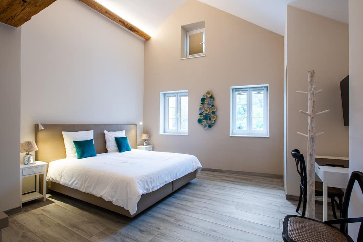 Un dormitorio con una cama grande, un escritorio y una silla. Hay dos ventanas y un techo con vigas a la vista.