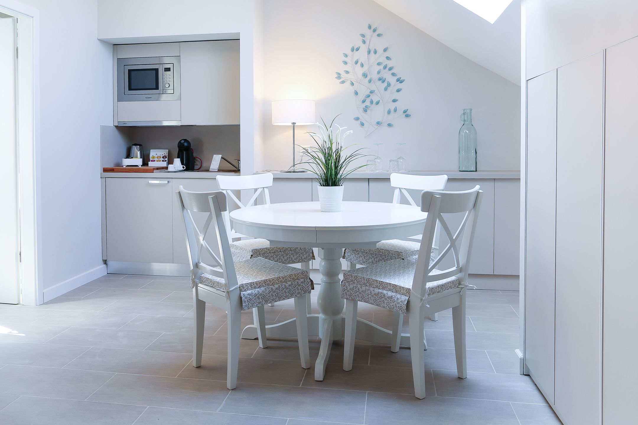 A modern kitchen with white cabinetry, a microwave, and a coffee maker on the counter. There is a dining table with four chairs and a potted plant in the center.