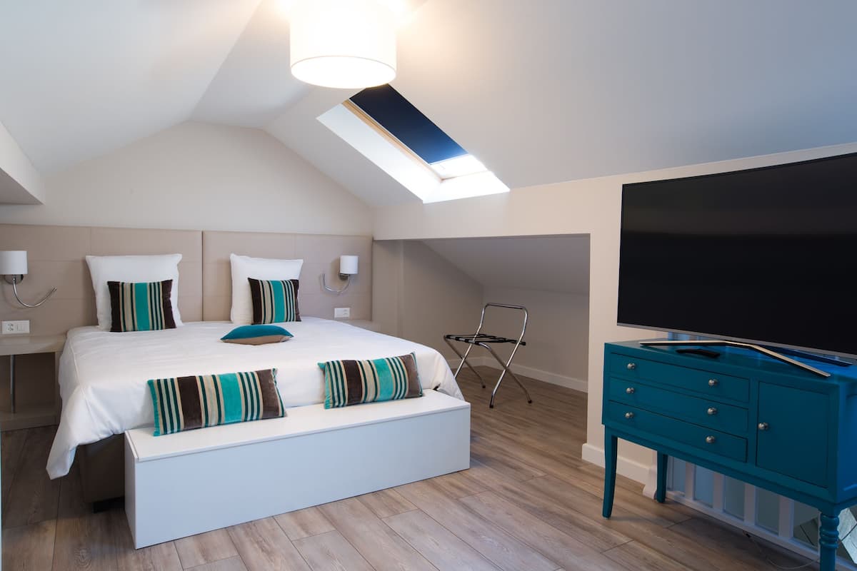 Un dormitorio con tragaluz, una cama con almohadas a rayas, un banco blanco, un aparador color turquesa y un televisor de pantalla plana.
