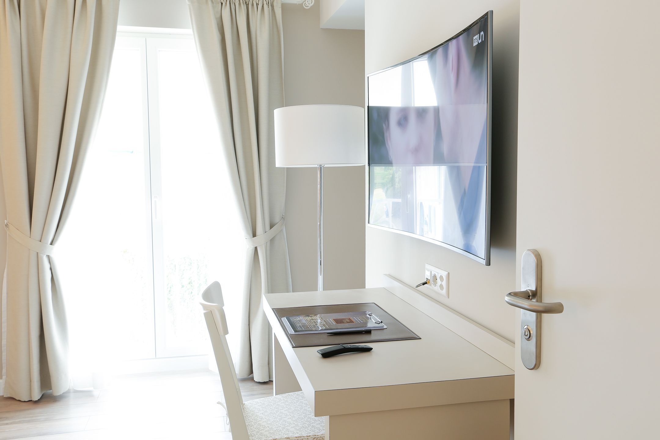 A hotel room with a desk, a flat-screen TV mounted on the wall, a lamp, and a door with a modern handle.