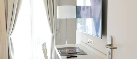 A hotel room with a desk, a flat-screen TV mounted on the wall, a lamp, and a door with a modern handle.