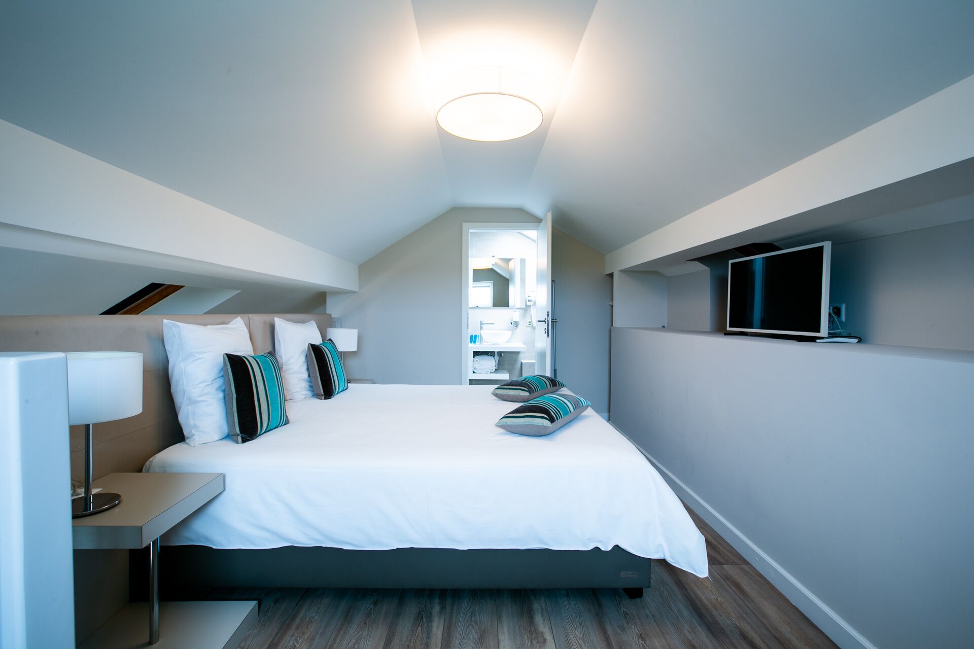 Una habitación de hotel moderna con una cama, mesita de noche, lámpara y televisor montado en la pared.