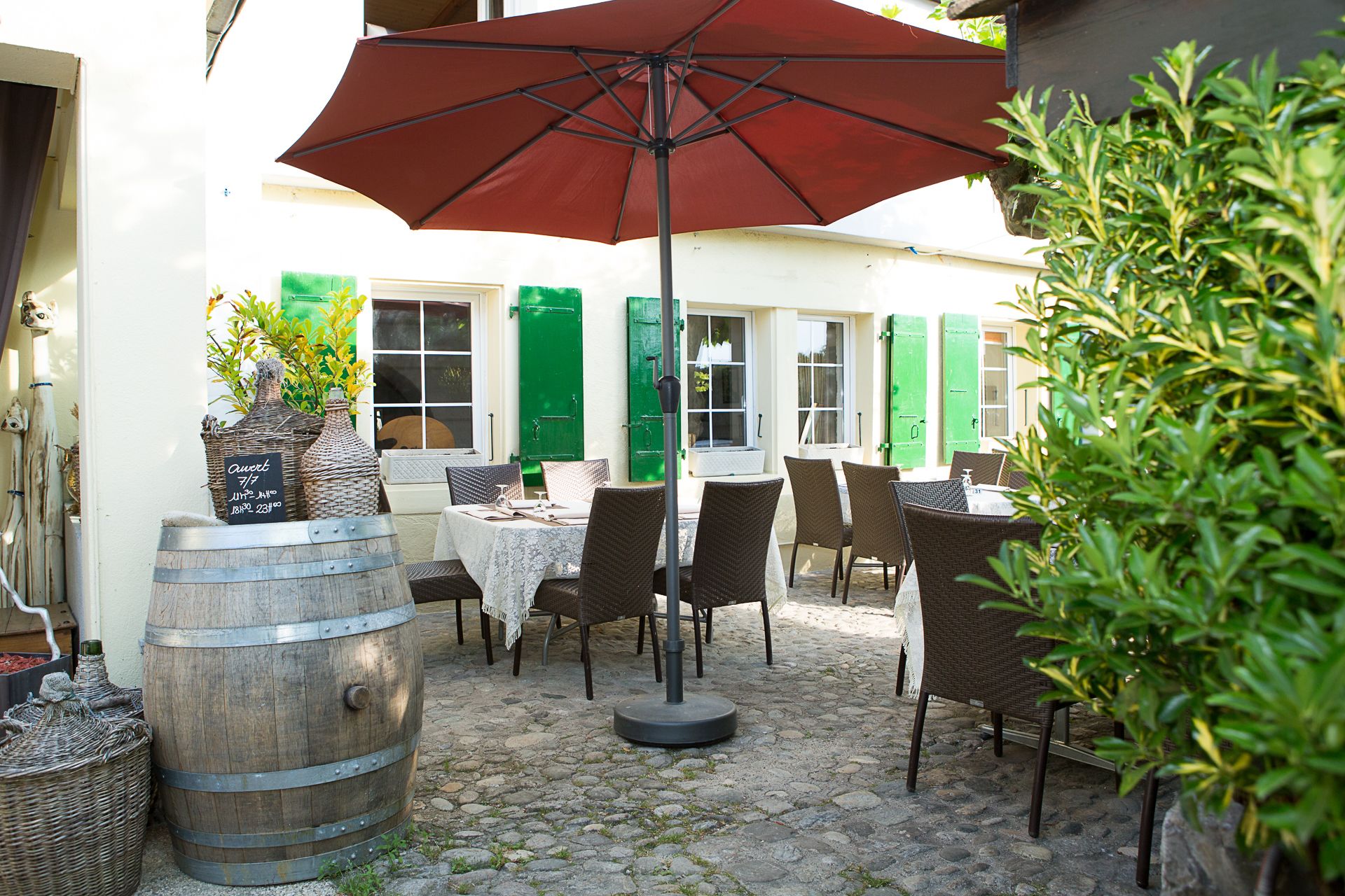 Een terras buiten met rieten meubels, een rode parasol en een stenen vloer.